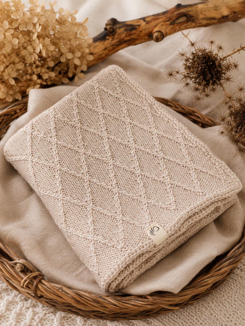 Handgestrickte Babydecke aus mulesingfreier Merinowolle in Naturbeige, dekorativ auf einem Leinentuch mit Holz, Trockenblumen und Herz in Szene gesetzt – nachhaltige Babydecke für Neugeborene und Geschenk zur Geburt.