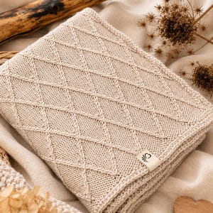 Handgestrickte Babydecke aus mulesingfreier Merinowolle in Naturbeige, dekorativ auf einem Leinentuch mit Holz, Trockenblumen und Herz in Szene gesetzt – nachhaltige Babydecke für Neugeborene und Geschenk zur Geburt.