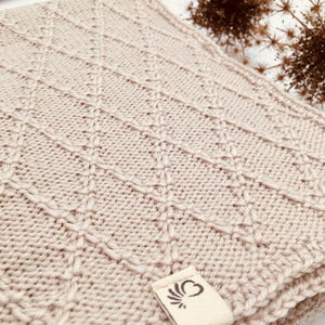 May include: A light beige knitted blanket featuring a raised diamond pattern across the surface. A small, rectangular off-white fabric tag is attached to the corner, displaying a dark brown graphic logo of a stylized heart with radiating lines.