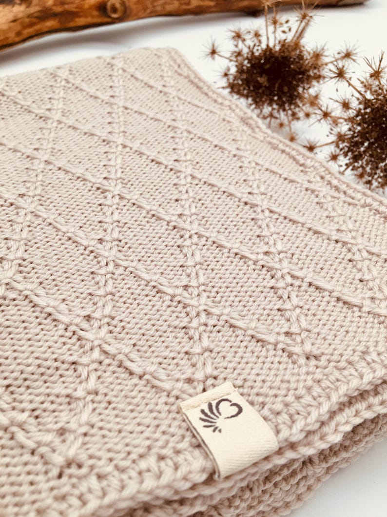 May include: A light beige knitted blanket featuring a raised diamond pattern across the surface. A small, rectangular off-white fabric tag is attached to the corner, displaying a dark brown graphic logo of a stylized heart with radiating lines.