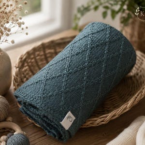 May include: A folded teal blue knit blanket featuring a raised diamond pattern across its surface. The blanket has a small white rectangular tag attached to one corner with a circular logo graphic printed in dark ink on the fabric surface.