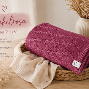 May include: A dark pink knitted blanket with a diamond pattern texture, folded inside a woven basket. The blanket features a small white rectangular tag with a heart logo. Beside it lies a beige crocheted rabbit toy and a light fabric cloth.