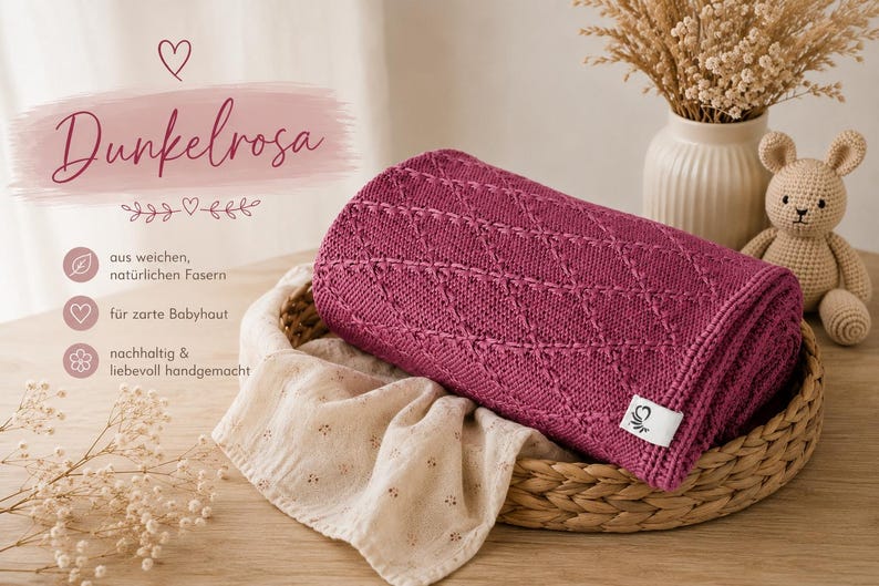 May include: A dark pink knitted blanket with a diamond pattern texture, folded inside a woven basket. The blanket features a small white rectangular tag with a heart logo. Beside it lies a beige crocheted rabbit toy and a light fabric cloth.