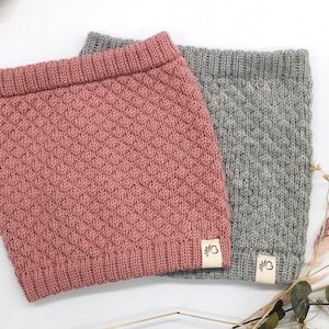 Könnte beinhalten: Zwei gestrickte Halstücher im Bild. Eines ist staubrosa, das andere grau. Beide haben ein strukturiertes, rautenförmiges Muster und ein kleines Etikett. Die Halstücher werden auf einer weißen Oberfläche präsentiert.