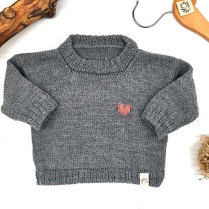 Può includere: Un maglione grigio lavorato a maglia con un piccolo cuore rosa sul petto. Il maglione ha un girocollo e maniche lunghe. Una piccola etichetta è cucita sul fondo. Il maglione è esposto su una superficie bianca con una gruccia di legno.
