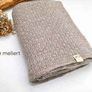 Handgestrickte Decke in Beige meliert mit feiner Strickstruktur, Joseda-Etikett, dekoriert mit Trockenblumen und Holzast auf weißem Hintergrund.