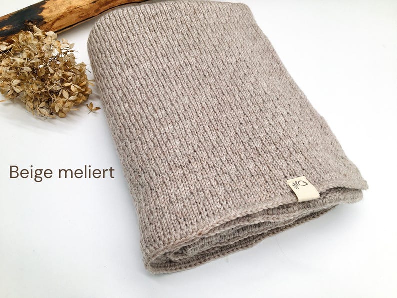 Handgestrickte Decke in Beige meliert mit feiner Strickstruktur, Joseda-Etikett, dekoriert mit Trockenblumen und Holzast auf weißem Hintergrund.