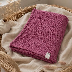 May include: A folded magenta knitted blanket featuring a textured diamond pattern across its surface. The blanket includes a small white rectangular fabric tag in the bottom right corner with a black graphic logo of a stylized flower or leaf.