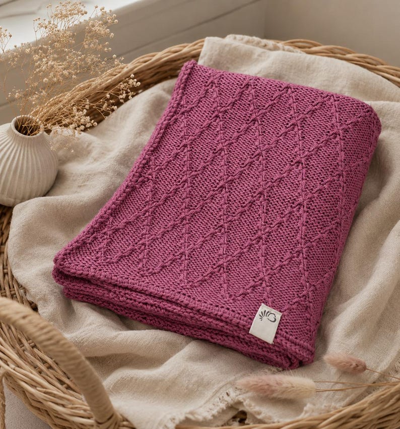 May include: A folded magenta knitted blanket featuring a textured diamond pattern across its surface. The blanket includes a small white rectangular fabric tag in the bottom right corner with a black graphic logo of a stylized flower or leaf.