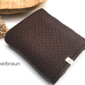 Handgestrickte Babydecke in Dunkelbraun mit feiner Strickstruktur, weiches Naturgarn mit Joseda-Label, dekoriert mit Trockenblumen und Holzschale auf hellem Hintergrund, Farbbezeichnung „Dunkelbraun“ im Bild.