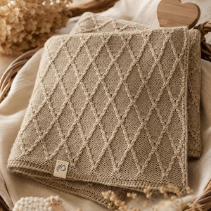 Handgestrickte Babydecke aus mulesingfreier Merinowolle in Naturbeige, dekorativ auf einem Leinentuch mit Trockenblumen, Holzherz und Schäfchen arrangiert – hochwertige, nachhaltige Babydecke für Neugeborene und Geschenk zur Geburt.