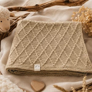 Handgestrickte Babydecke aus mulesingfreier Merinowolle in Naturbeige, dekorativ auf einem Leinentuch mit Trockenblumen, Holzherz und Schäfchen arrangiert – hochwertige, nachhaltige Babydecke für Neugeborene und Geschenk zur Geburt.
