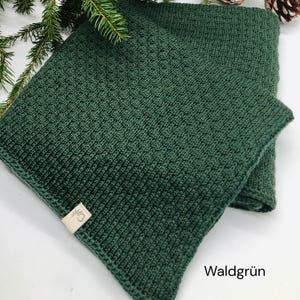 Handgestrickte Babydecke in Waldgrün mit feiner Strickstruktur, weiches Naturgarn mit Joseda-Label, dekoriert mit Tannenzweigen und Zapfen auf hellem Hintergrund, Farbbezeichnung „Waldgrün“ im Bild.