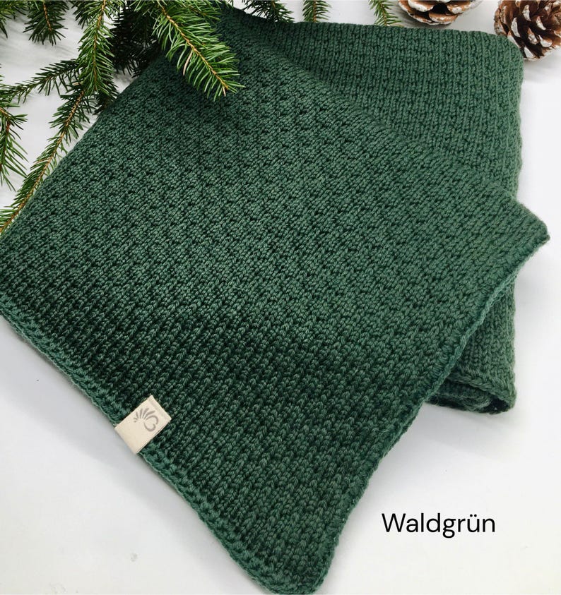 Handgestrickte Babydecke in Waldgrün mit feiner Strickstruktur, weiches Naturgarn mit Joseda-Label, dekoriert mit Tannenzweigen und Zapfen auf hellem Hintergrund, Farbbezeichnung „Waldgrün“ im Bild.