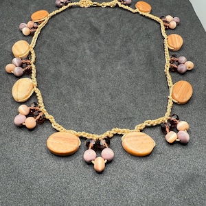 Puede incluir: Un collar dorado con un diseño de cadena trenzada. El collar está adornado con piedras grandes y redondas de color melocotón y racimos de cuentas más pequeñas, redondas, rosas y marrones. El collar se muestra sobre un fondo gris oscuro.