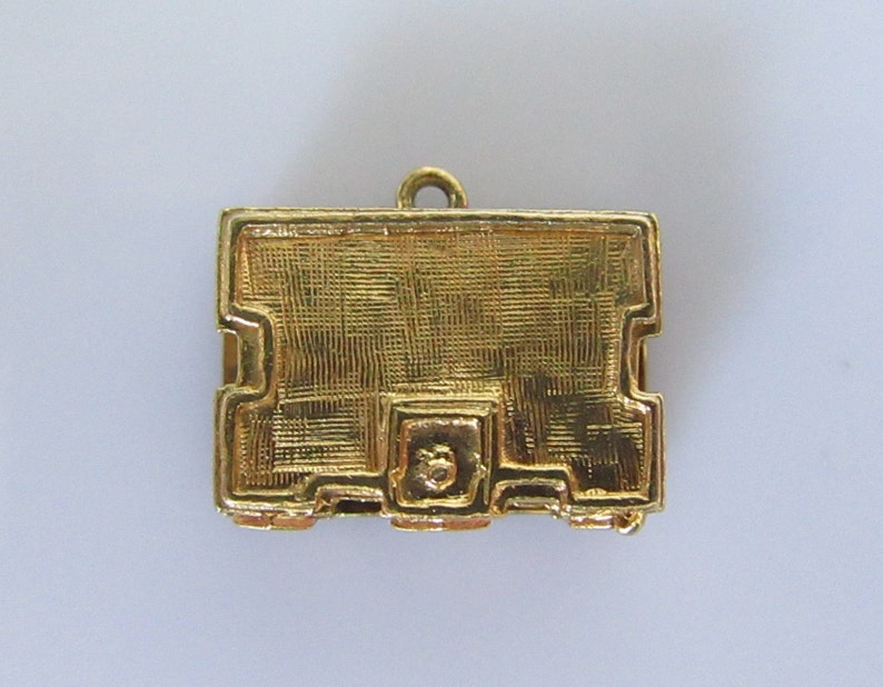 Puede incluir: Un amuleto de metal dorado en forma de peque&ntilde;a caja rectangular con una superficie texturizada. El amuleto tiene un bucle en la parte superior para sujetarlo a un collar o pulsera.