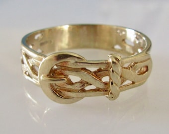 May include: A gold-toned ring with a detailed, intricate design of a belt buckle. The ring is made of metal and has a polished finish.