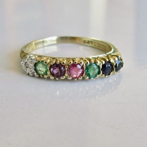 May include: A gold band ring featuring a row of seven round faceted gemstones in various colors including green, purple, pink, and dark blue, alongside a small cluster of clear white stones set into the yellow gold metal band.
