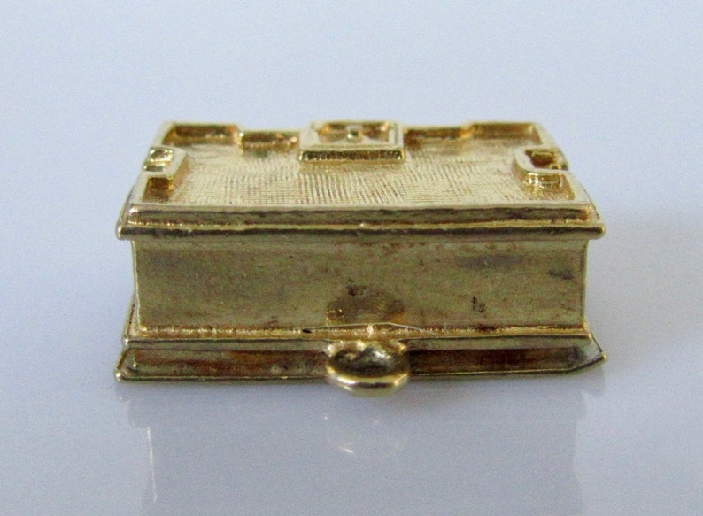 Puede incluir: Un amuleto de metal dorado en forma de libro cerrado con un peque&ntilde;o lazo en la parte inferior.
