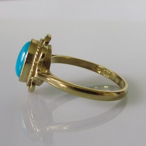 May include: A gold ring with a turquoise gemstone set in a decorative design. The ring is on a white surface.