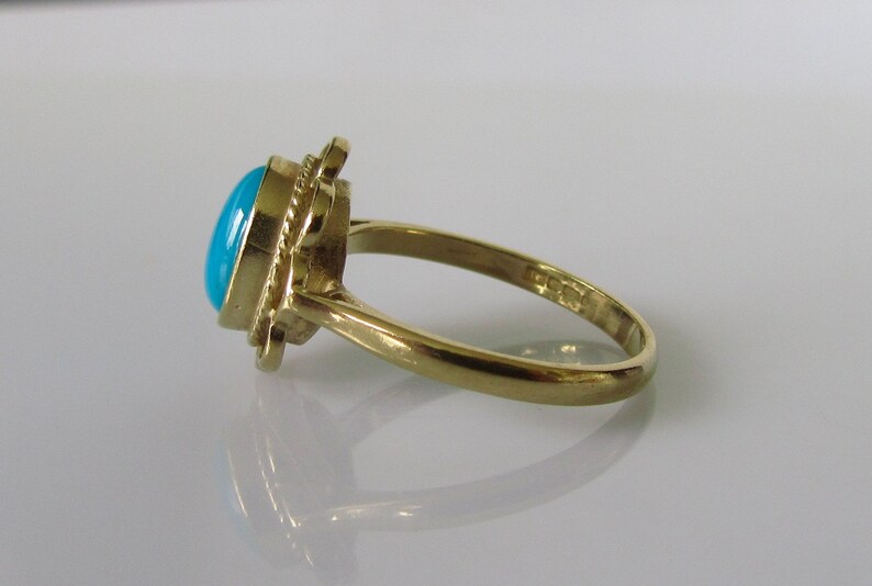 May include: A gold ring with a turquoise gemstone set in a decorative design. The ring is on a white surface.