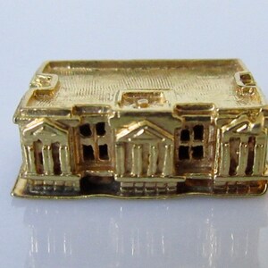 Puede incluir: Un amuleto de edificio en miniatura de color dorado con una fachada detallada y un techo plano. El edificio tiene un dise&ntilde;o sim&eacute;trico con columnas y ventanas.