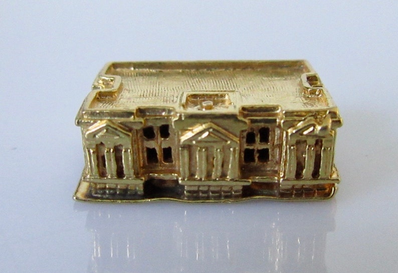 Puede incluir: Un amuleto de edificio en miniatura de color dorado con una fachada detallada y un techo plano. El edificio tiene un dise&ntilde;o sim&eacute;trico con columnas y ventanas.