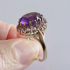May include: A gold ring with a large, oval-shaped amethyst gemstone surrounded by a halo of smaller amethyst stones.