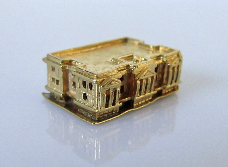Puede incluir: Un amuleto de oro con forma de edificio con una fachada detallada. El amuleto est&aacute; hecho de oro macizo y tiene un acabado brillante.