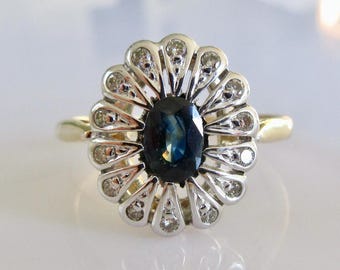 May include: A gold ring with a blue sapphire center stone surrounded by a halo of small diamonds. The ring has a flower-shaped design.