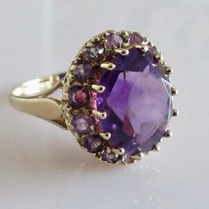 May include: A gold ring with a large, faceted amethyst gemstone surrounded by a halo of smaller amethyst stones.