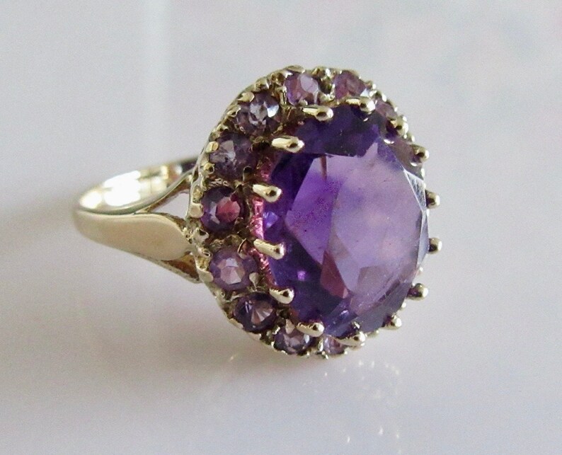 May include: A gold ring with a large, faceted amethyst gemstone surrounded by a halo of smaller amethyst stones.