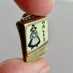 Pu&ograve; includere: Un ciondolo d'oro con sfondo smaltato bianco e un'illustrazione in bianco e nero di una donna con un abito tradizionale gallese. Il ciondolo &egrave; inciso con la parola "WALES".