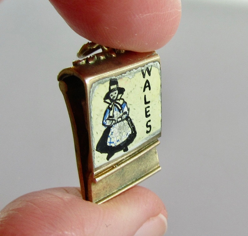 Pu&ograve; includere: Un ciondolo d'oro con sfondo smaltato bianco e un'illustrazione in bianco e nero di una donna con un abito tradizionale gallese. Il ciondolo &egrave; inciso con la parola "WALES".