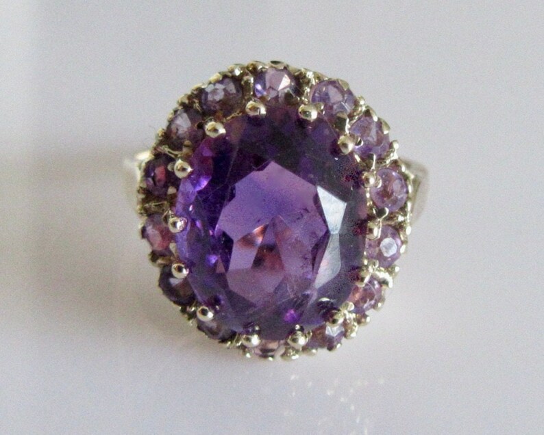 May include: A silver ring with a large, oval-shaped amethyst gemstone surrounded by a halo of smaller amethyst stones.