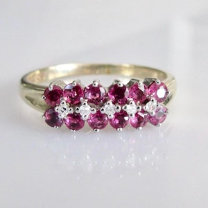 Pode incluir: Um anel dourado com duas fileiras de pedras preciosas redondas rosa escuro, intercaladas com pedras preciosas claras menores. A banda do anel é lisa e polida. Uma joia.