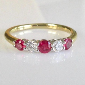 Puede incluir: Un anillo de oro con cinco piedras preciosas. Alterna rubíes rojos de talla ovalada y diamantes redondos y claros. El anillo está colocado sobre una superficie blanca que refleja la luz.