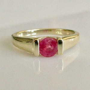 May include: A gold ring with a round, faceted ruby gemstone. The ring has a simple, elegant design with a polished finish. The ruby is a vibrant pink-red color, set in a classic bezel setting.