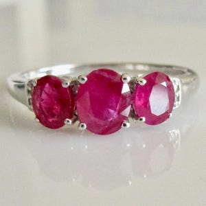 May include: A silver ring with three oval-shaped ruby gemstones. The rubies are a deep red color and are set in a prong setting.