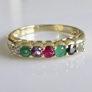 Puede incluir: Un anillo de oro con una serie de piedras preciosas de colores. El anillo presenta una fila de esmeraldas, rubíes y otras piedras preciosas. El anillo está sobre una superficie blanca.