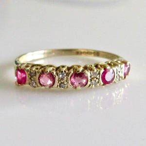 May include: A gold ring with a row of pink gemstones and small white stones. The ring's band is a warm gold colour, and the gemstones are set in individual, ornate settings. The ring is displayed on a white surface.
