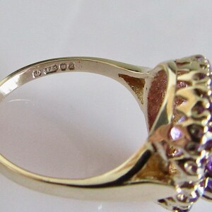 May include: A gold ring with a large purple amethyst gemstone surrounded by smaller amethyst stones. The ring is marked with the number "925" on the inside band.