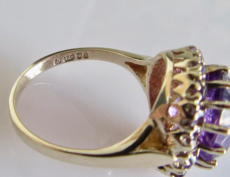 May include: A gold ring with a large purple amethyst gemstone surrounded by smaller amethyst stones. The ring is marked with the number "925" on the inside band.