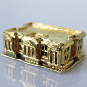 Puede incluir: Un amuleto de edificio en miniatura de color dorado con detalles intrincados, que se asemeja a una estructura arquitect&oacute;nica cl&aacute;sica.