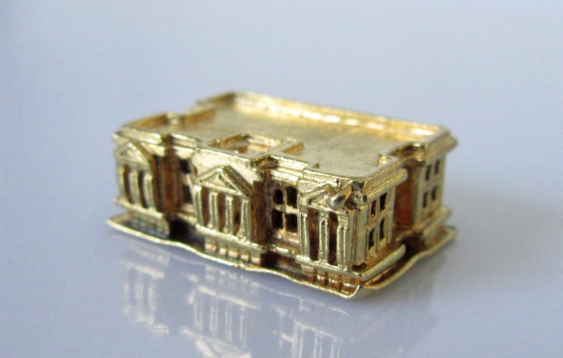 Puede incluir: Un amuleto de edificio en miniatura de color dorado con detalles intrincados, que se asemeja a una estructura arquitect&oacute;nica cl&aacute;sica.