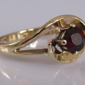 May include: A gold ring with a swirling design and a single red gemstone in the center.