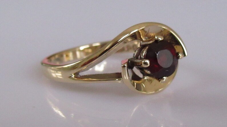 May include: A gold ring with a swirling design and a single red gemstone in the center.