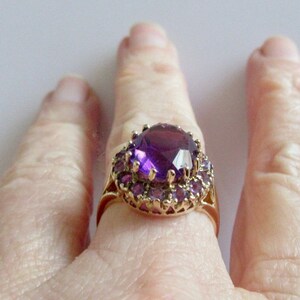 May include: A gold ring with a large, faceted, purple amethyst gemstone surrounded by a halo of smaller purple amethysts.