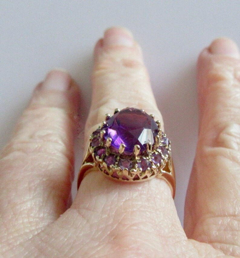 May include: A gold ring with a large, faceted, purple amethyst gemstone surrounded by a halo of smaller purple amethysts.