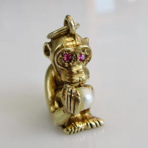 Puede incluir: Un dije de mono dorado con ojos de rubí y una perla en sus manos. El dije está detallado con pelaje texturizado y un lazo para sujetarlo a un collar o pulsera.
